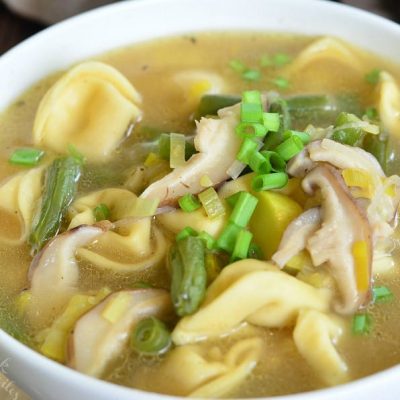 White bowl filled with vegetable medley tortellini soup on a wooden table with a spoon to the left and a tan cloth in the background as viewed close up