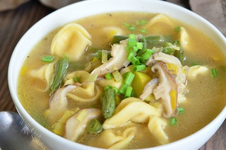 White bowl filled with vegetable medley tortellini soup on a wooden table with a spoon to the left and a tan cloth in the background as viewed close up