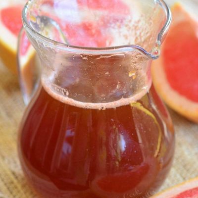 Glass jar filled with homemade grapefruit syrup and sauce on a tan placemat with a wooden spoon leaning on the jar and sliced grapefruit laying around the jar.