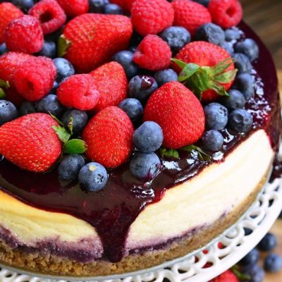 Whole mixed berry cheesecake on a cake platter with berries below the platter as well as viewed close up