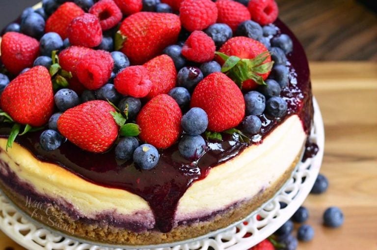 Whole mixed berry cheesecake on a cake platter with berries below the platter as well as viewed close up