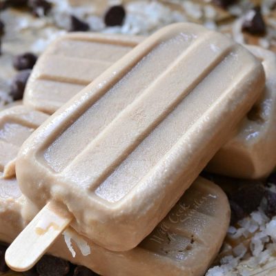 3 creamy coconut mocha ice pops stacked on each other while sitting on a wooden slatted table with coconut and chocolate chips scattered around the table