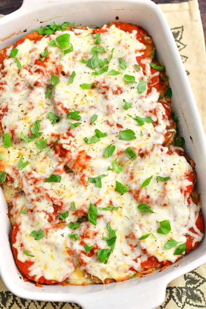 baked eggplant parmesan in a white casserole dish topped with fresh basil leaves and the dish is standing on a burlap towel