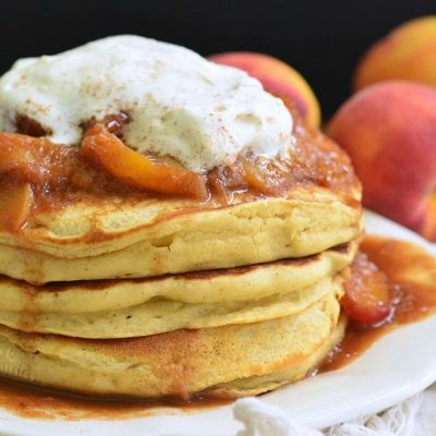 Stack of peaches and cream fluffy buttermilk pancakes on a white decorative plate with peaches in the background as viewed close up