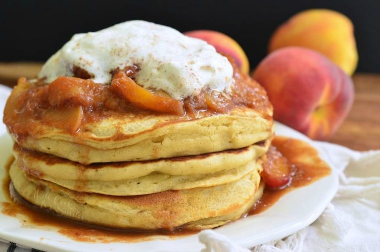 Stack of peaches and cream fluffy buttermilk pancakes on a white decorative plate with peaches in the background as viewed close up