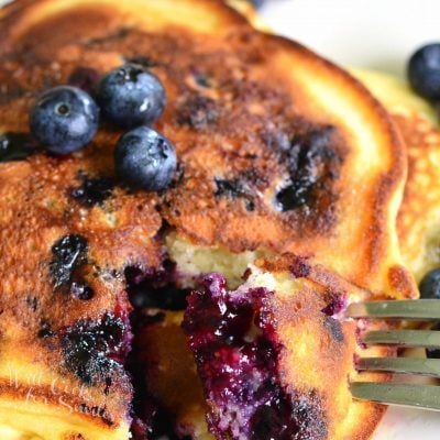 Single serving buttermilk pancakestopped with syrup and blueberries on a white plate with a taking one bite out of the pancakes.