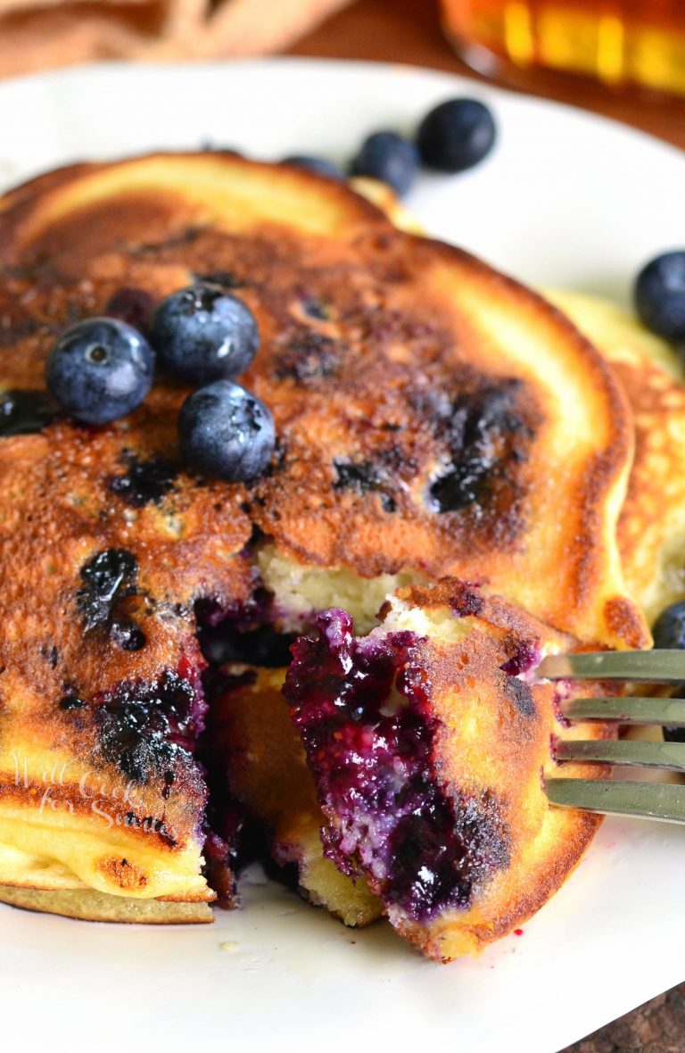 Single serving buttermilk pancakestopped with syrup and blueberries on a white plate with a taking one bite out of the pancakes.