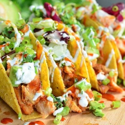 sweet and spicey siracha chicken tacos on a wooden cutting board with limes and avocado halves around them viewed close up