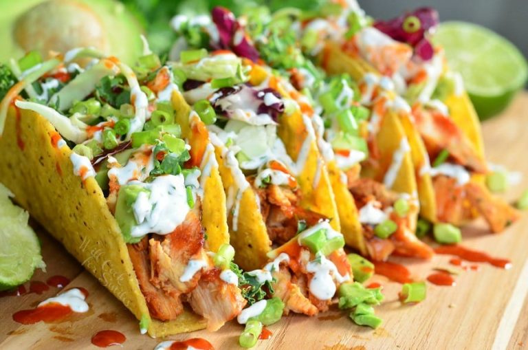 sweet and spicey siracha chicken tacos on a wooden cutting board with limes and avocado halves around them viewed close up