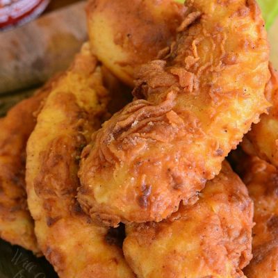 Siracha crisp chicken chicken tenders stacked in a pile with honey dipping sauce in a white bowl in the background.