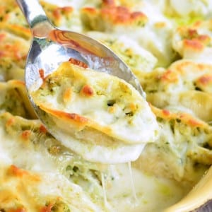 Cream colored baking pan with cheesy pesto chicken stuffed shells as viewd from slightly above with a serving spoon holding one shell above the rest.