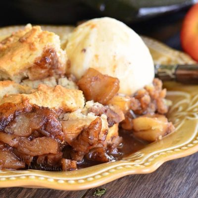 Large black skillet filled with maple pecan apple cobbler on a wooden table in the background with one portion on a decorative tan plate and topped with ice cream.