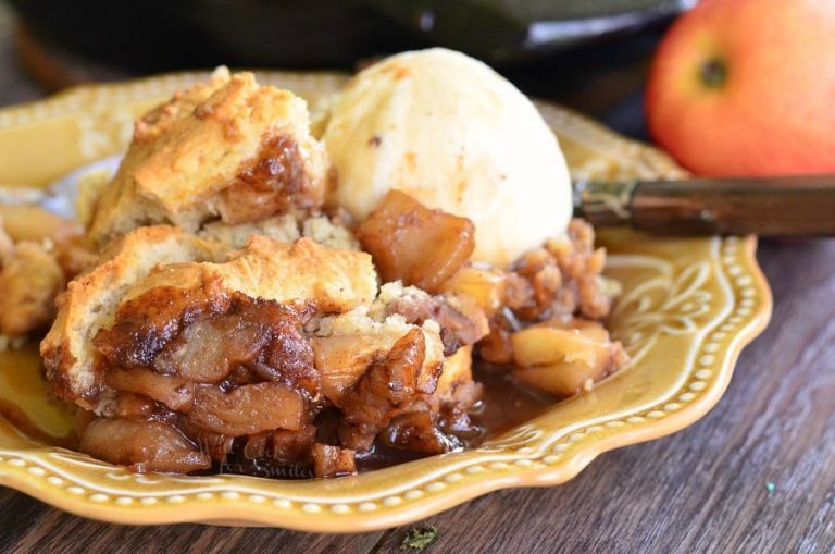 Large black skillet filled with maple pecan apple cobbler on a wooden table in the background with one portion on a decorative tan plate and topped with ice cream.