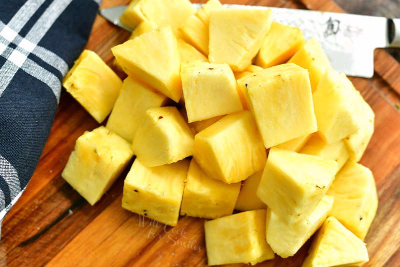 horizontal view of cut pineapple chunks in a mount on a cutting board with a knife next to it and a black plaid towel