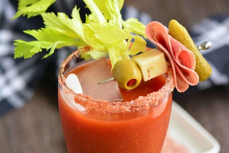 Top of glass shown filled with bloody mary and topped with a speared olive, pickle, cheese and meat garnish as a celery stalk rests on the rim of the glass. A wooden table below with a black and white cloth in the background.