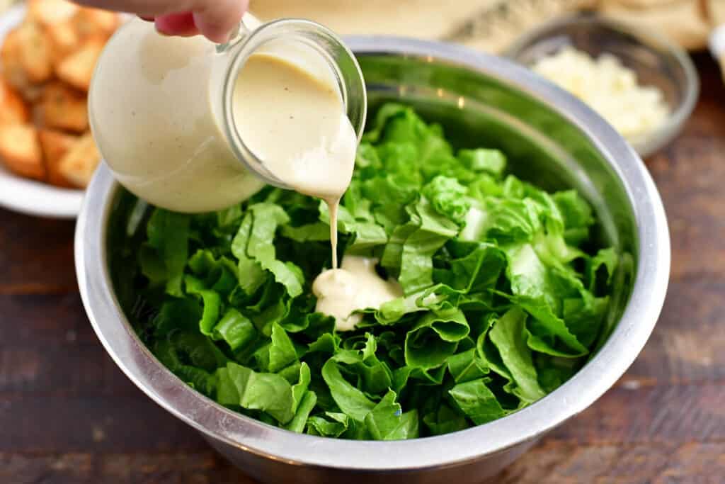 pouring white salad dressing over romaine lettuce leaves in salad bowl