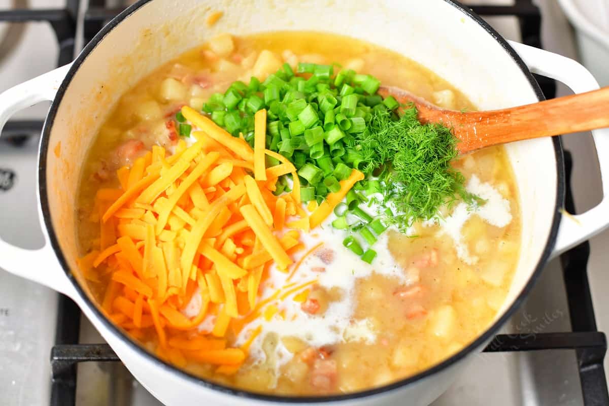 wooden spoon stirring shredded cheese, cream, dill, and green onion into a pot of soup on the stove