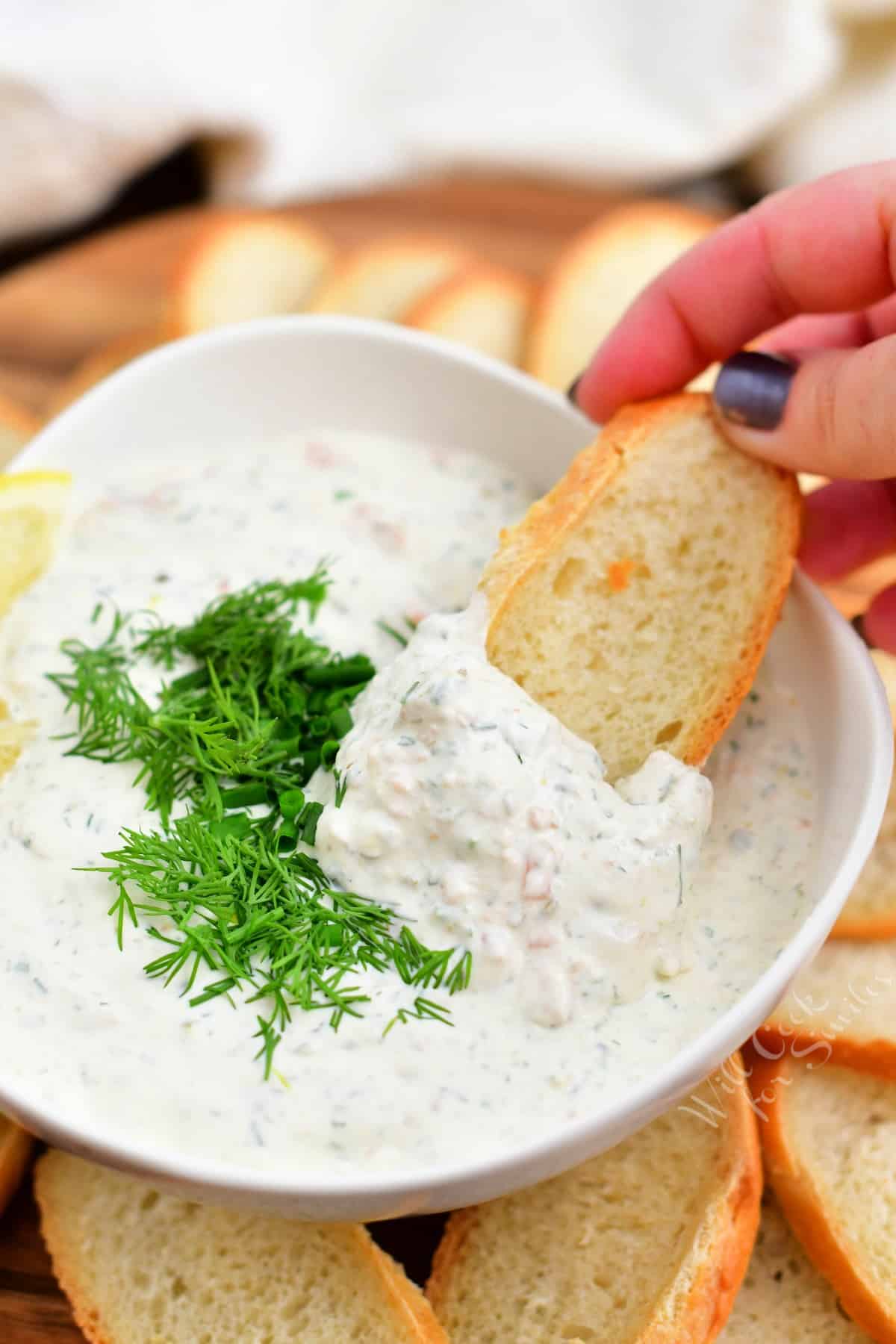 woman's hand dipping toasted baguette into bowl of creamy salmon dill dip