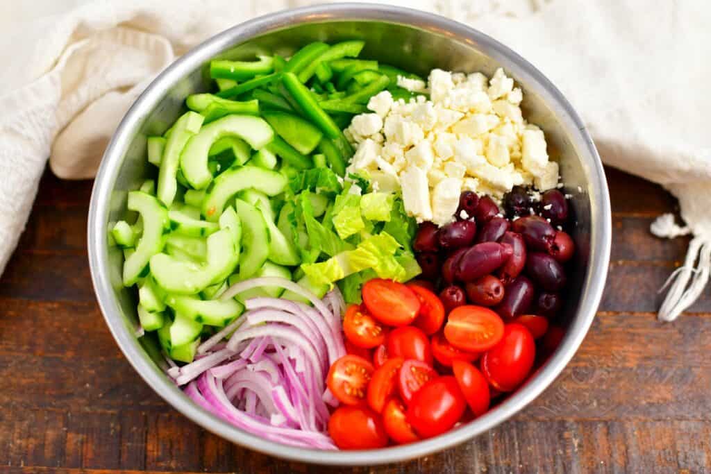 The ingredients for greek salad are all placed in a large silver mixing bowl.