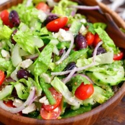 A freshly made greek salad is in a wooden bowl with a large wooden serving spoon.