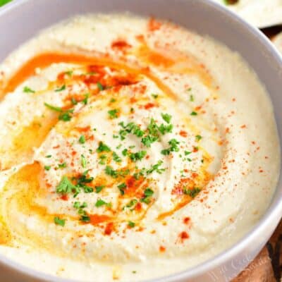 A white bowl filled with hummus is ready to be eaten.