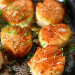 six visible seared scallops in a skillet with some parsley sprinkled on top.