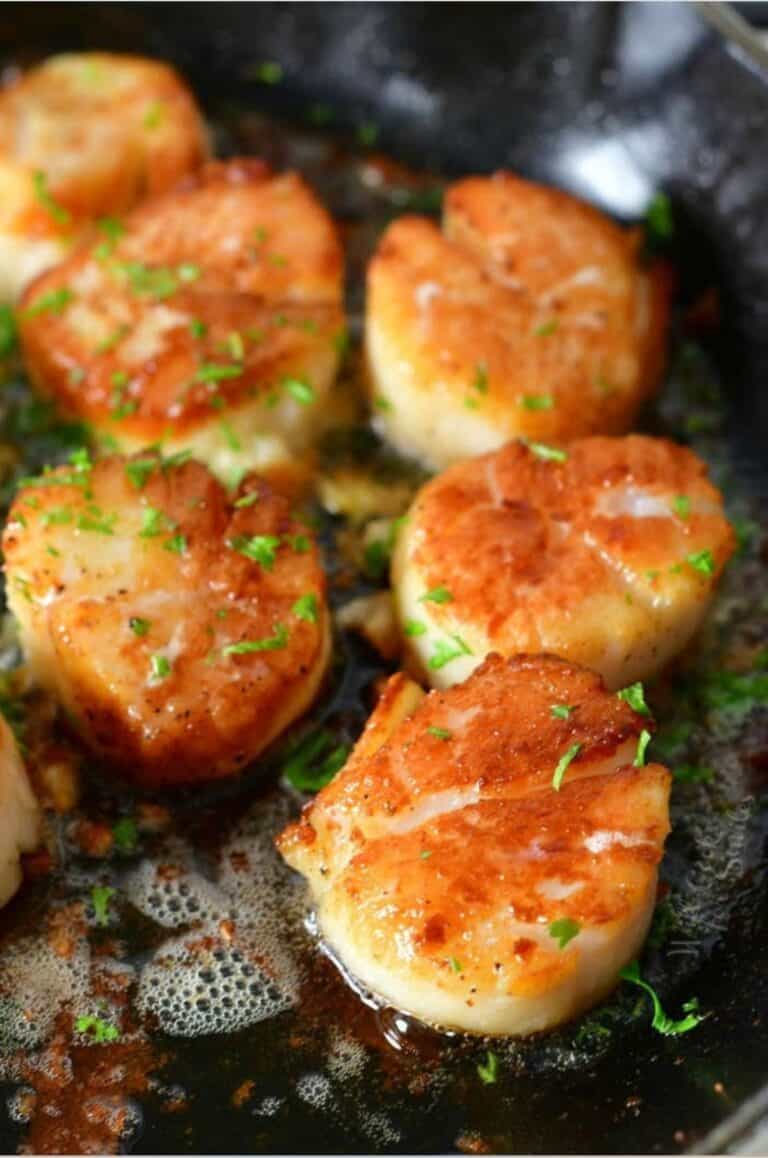 six visible seared scallops in a skillet with some parsley sprinkled on top.