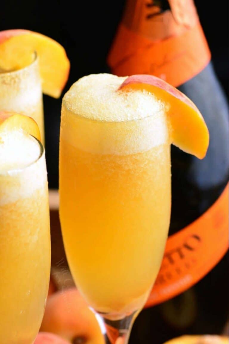 a champagne flute filled with orange peach Bellini cocktail with more on background.