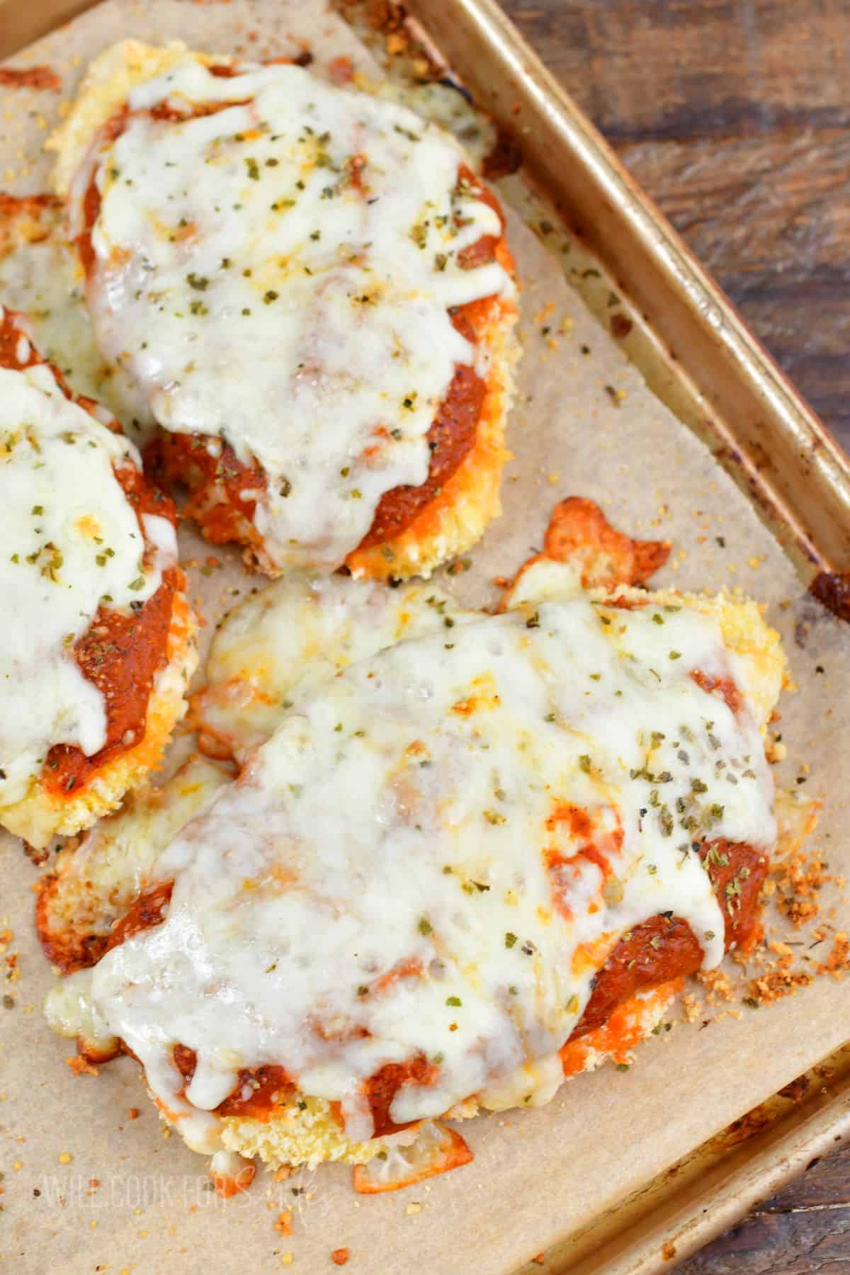 Three pieces of baked chicken parmesan on the baking sheet with melted cheese on top.