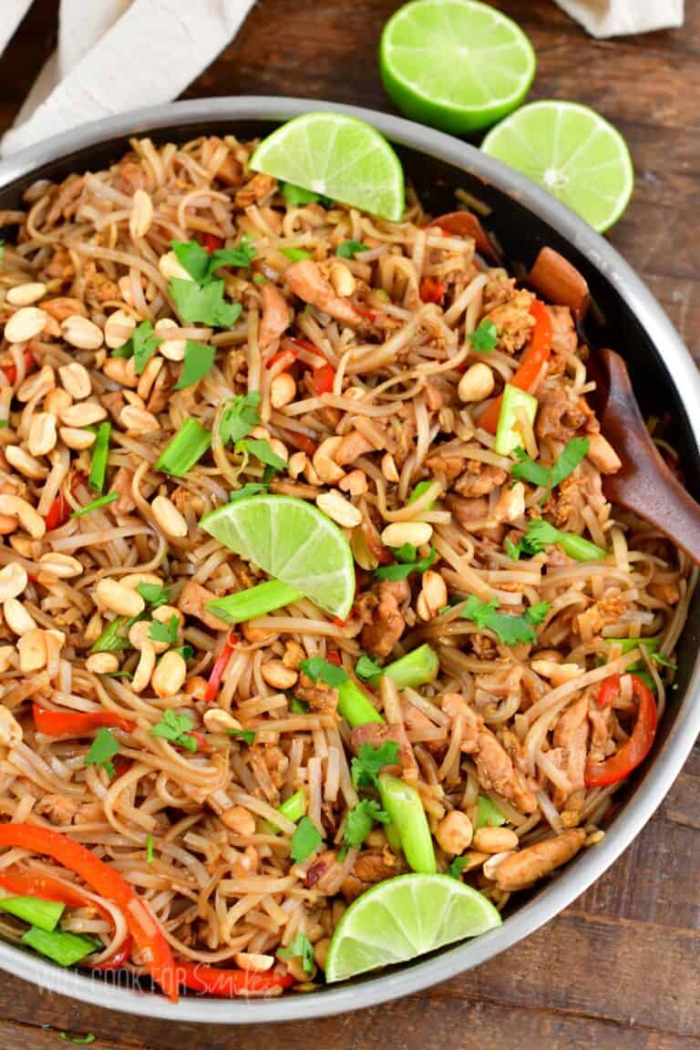 pad thai in a pan with lime wedges on a wood surface.