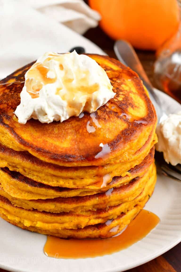 A stack of pumpkin pancakes with whipped cream and syrup on top on a plate.