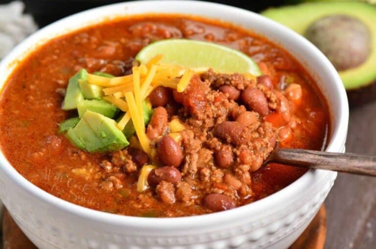 chili in a white bowl with a spoon in and cheese, avocado, and lime on top.