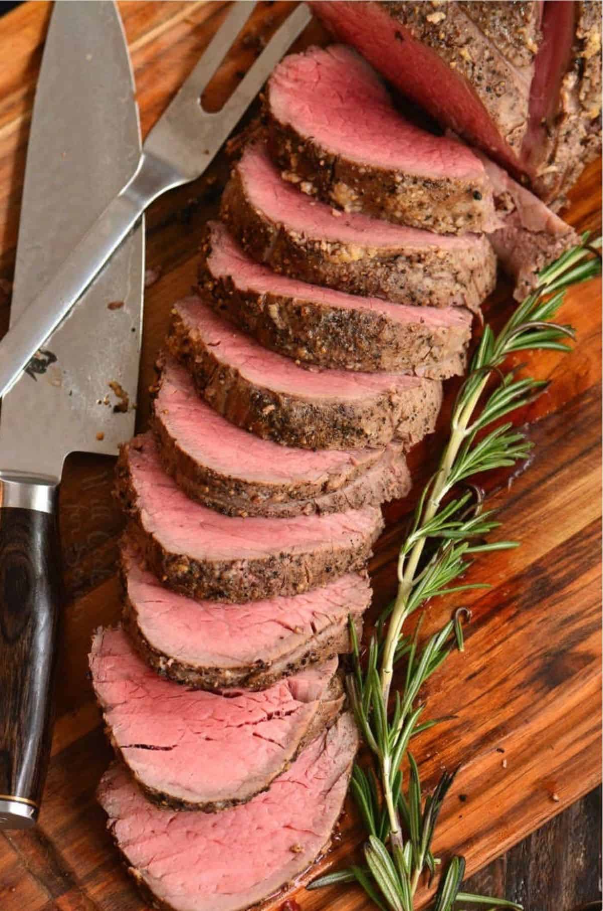 sliced beef tenderloin spread on cutting board next to a knife and rosemary.