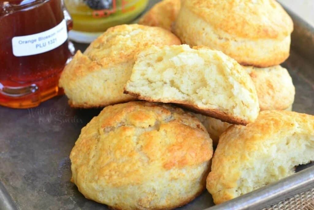 A tray of golden, freshly baked biscuits with one biscuit broken in half, revealing a fluffy interior. A jar of orange blossom honey is visible in the background.