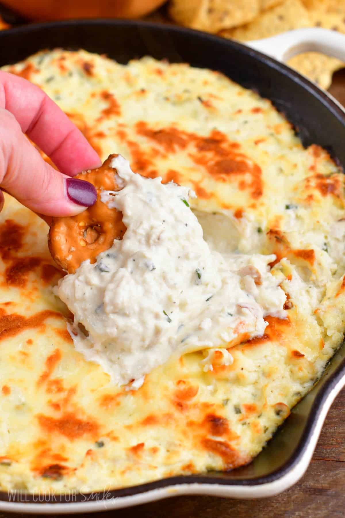 scooping out some creamy chicken ranch dip from the skillet with a pretzel chip.