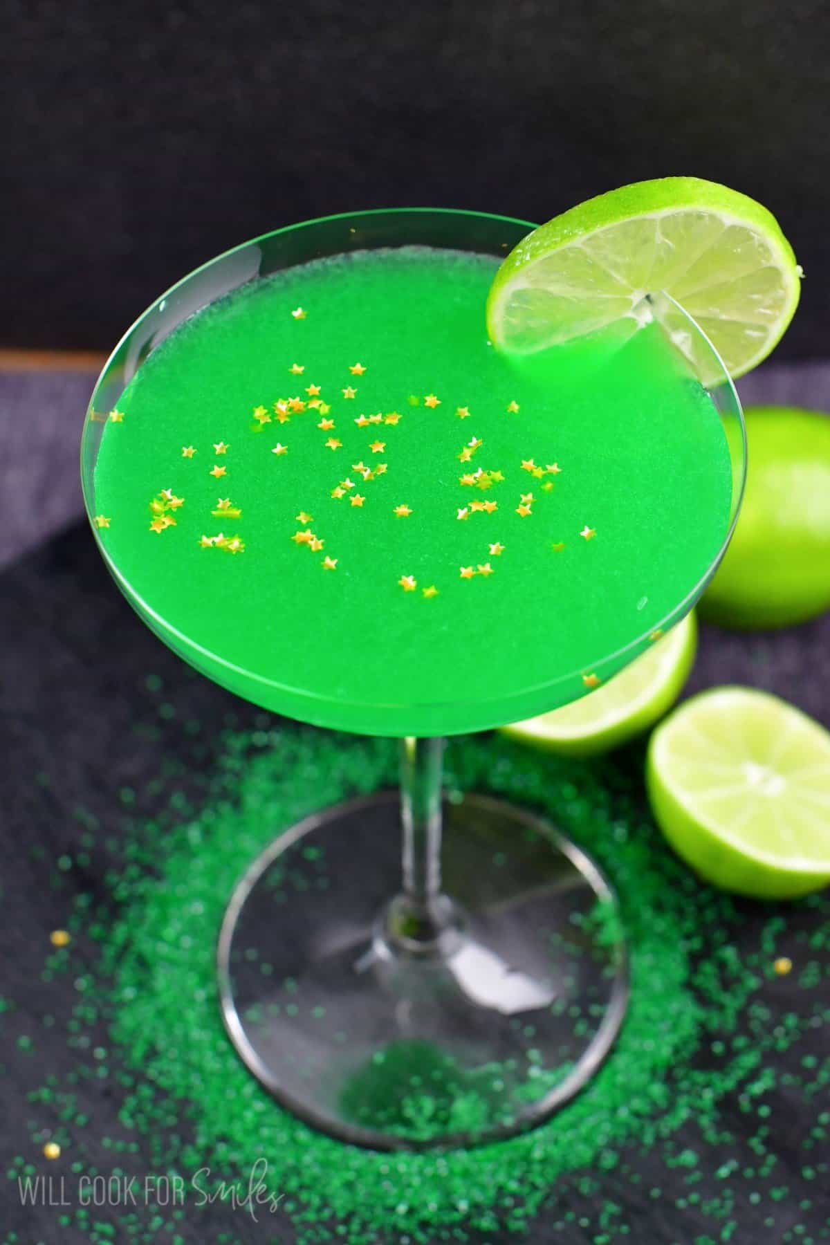 green martini in a martini glass with a lime on the edge and star sprinkles on top.