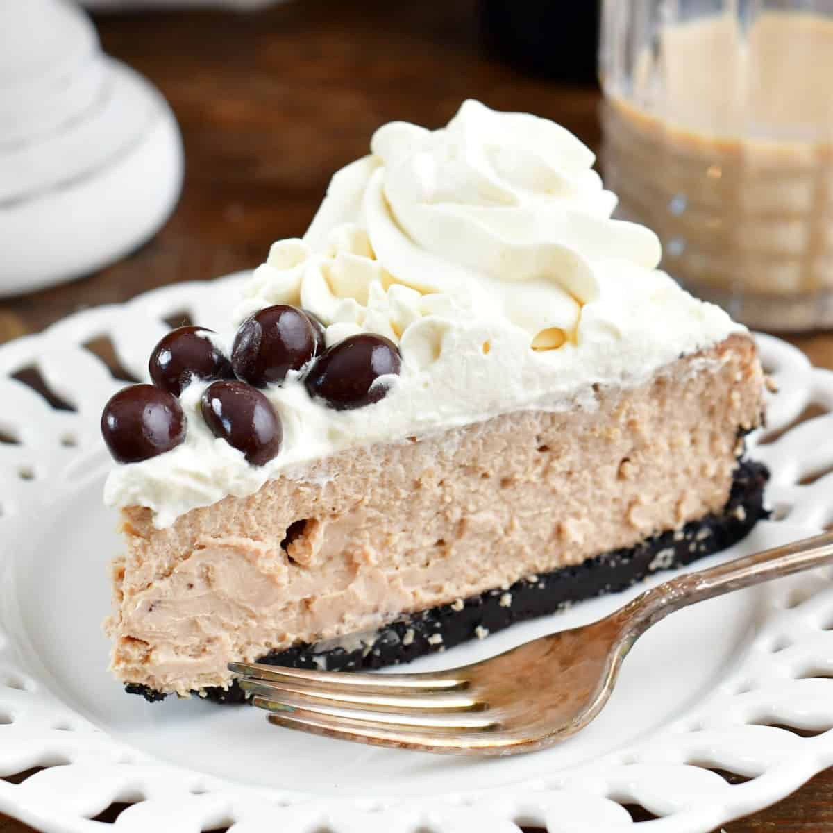 a slice of Baileys cheesecake on a white plate with a fork and some Irish cream in a glass.