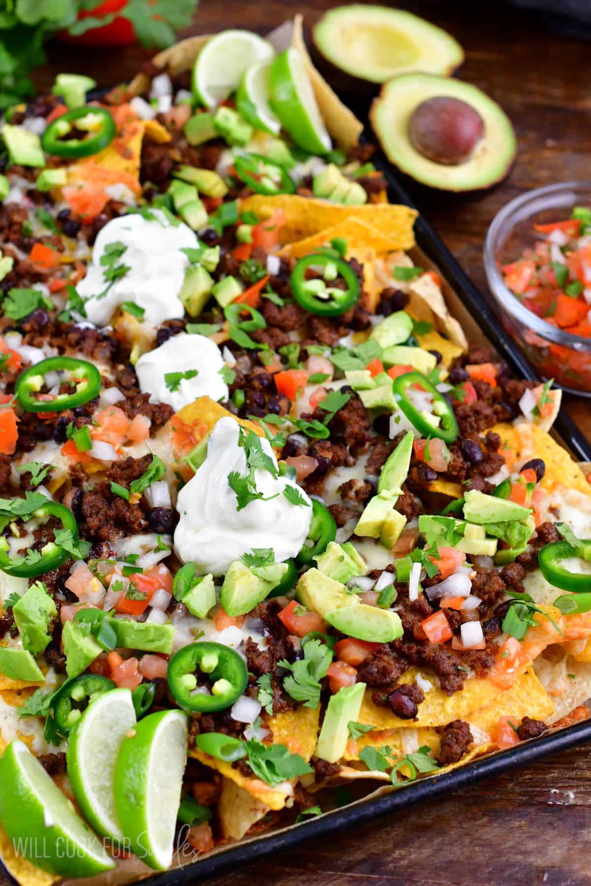 Beef nachos on a baking sheet with sour cream, jalapenos, onions, and lime wedges.