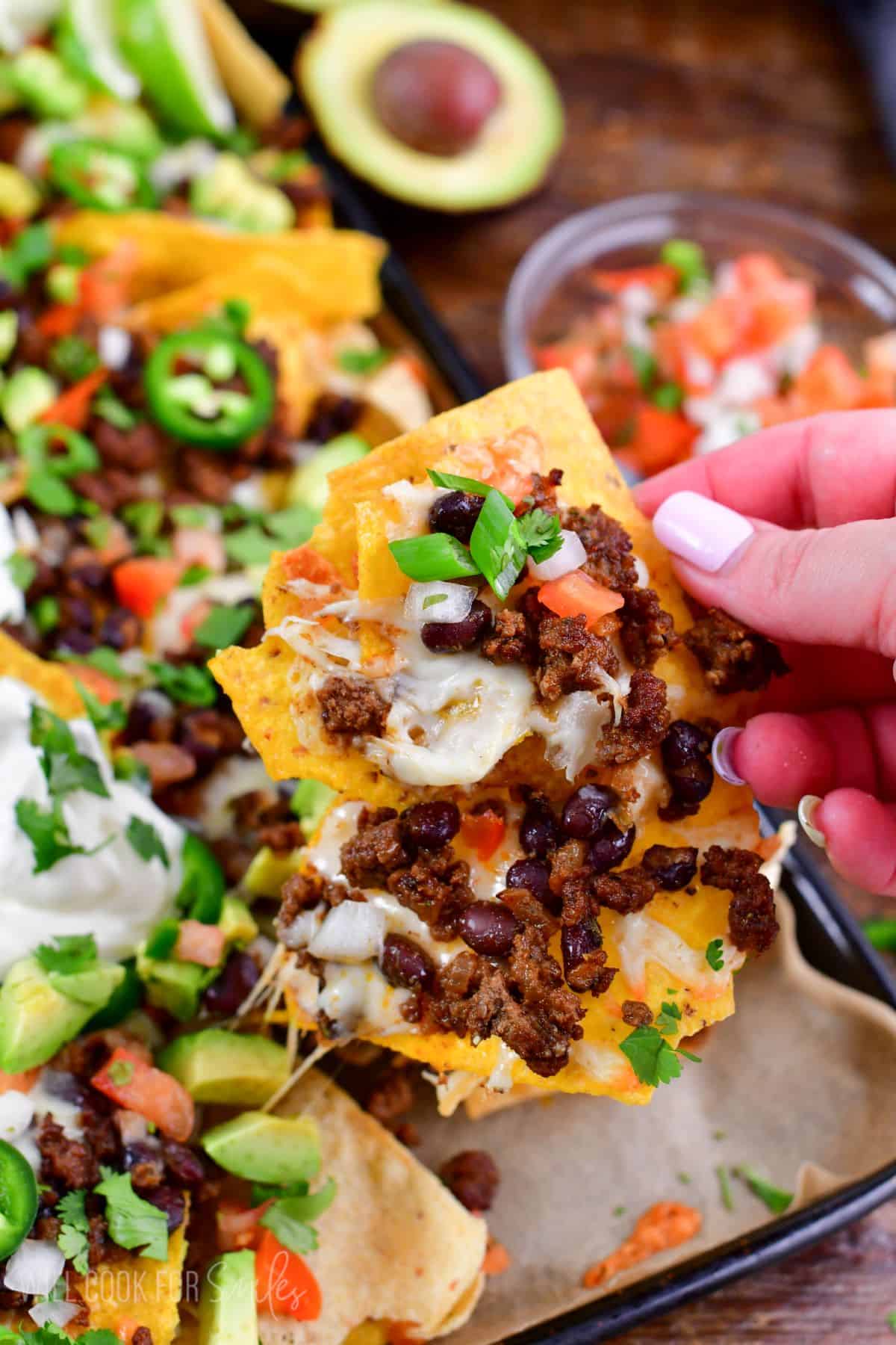 holding a beef nacho and the rest of the nachos in the background on a baking sheet.