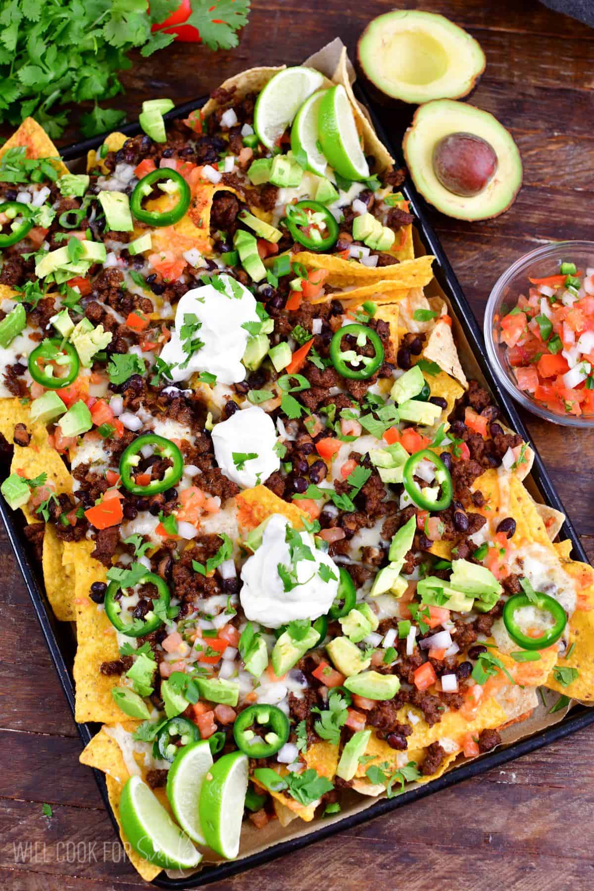 Beef nachos with jalapenos, avocado, sour cream, and limes on a baking sheet.