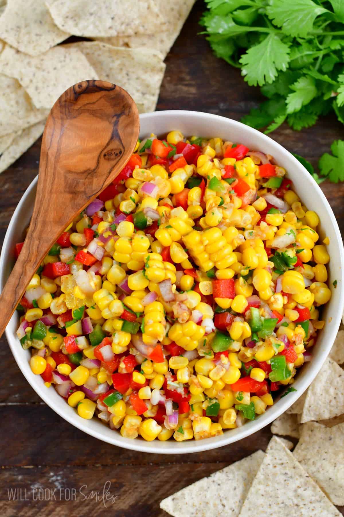 Corn salsa mixed up in a white bowl with a wooden spoon sitting over the bowl to the left.