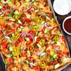 pulled pork nachos on baking sheet topped with pickled jalapenos, tomato, red onion, and diced avocado.