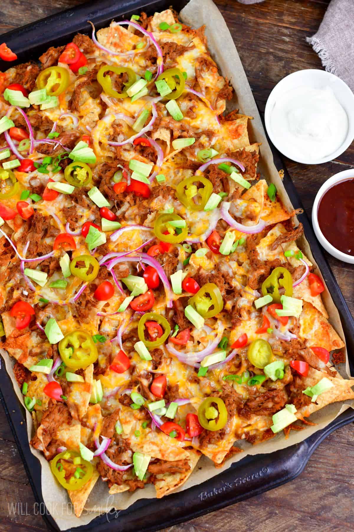 pulled pork nachos on baking sheet topped with pickled jalapenos, tomato, red onion, and diced avocado.