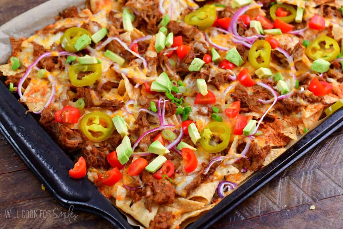 pulled pork nachos on a baking sheet topped with pickled jalapenos, tomatoes, red onion, and avocado.