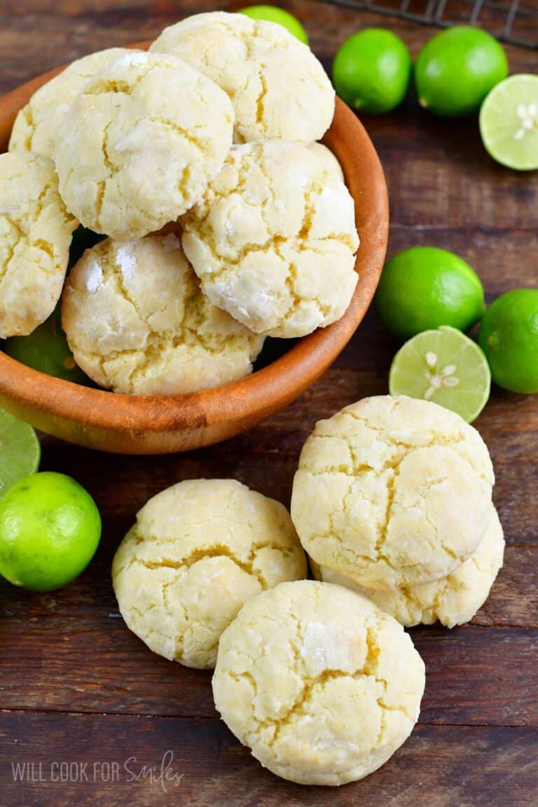 Cream cheese Cookies stacked in a wood bowl with a stack of cookies and limes beside the bowl on a wood surface.