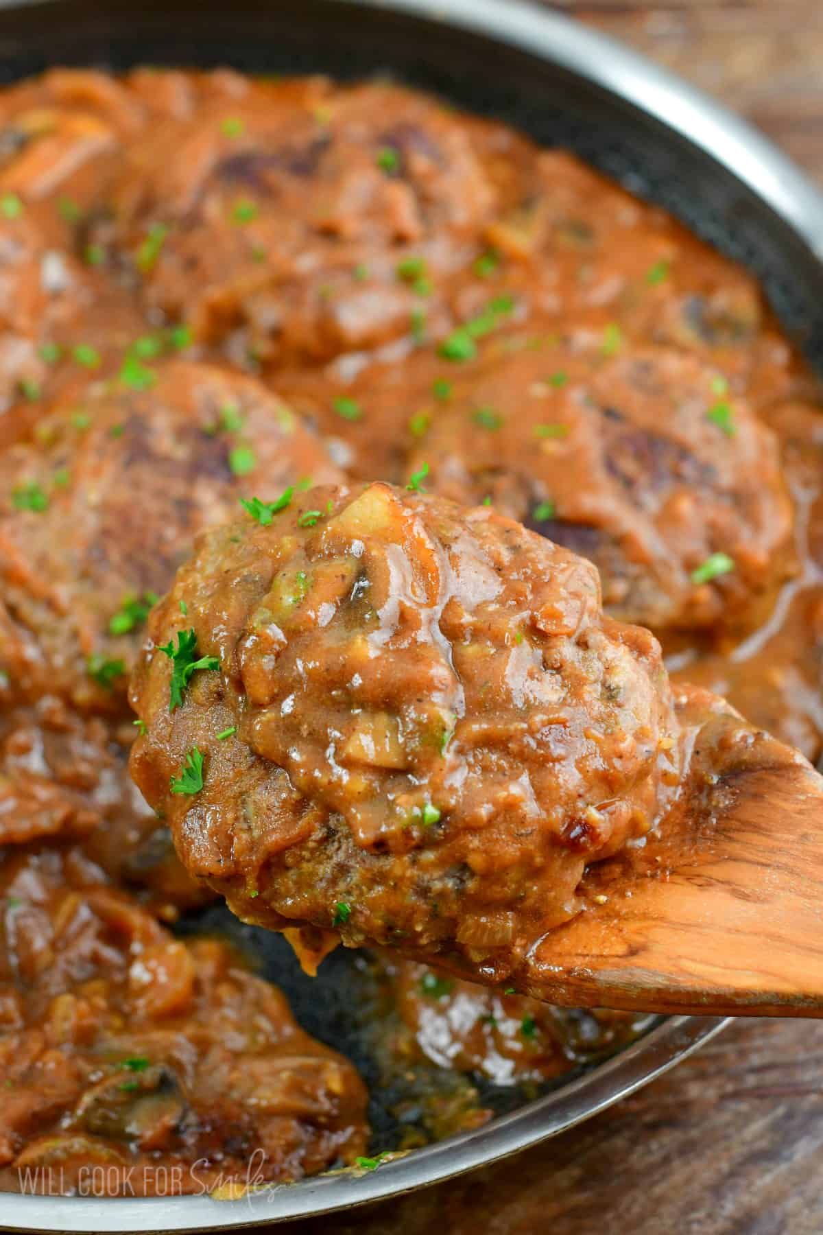 Holding a Salisbury steak on a wood spatula with the rest of the steaks in a pan.