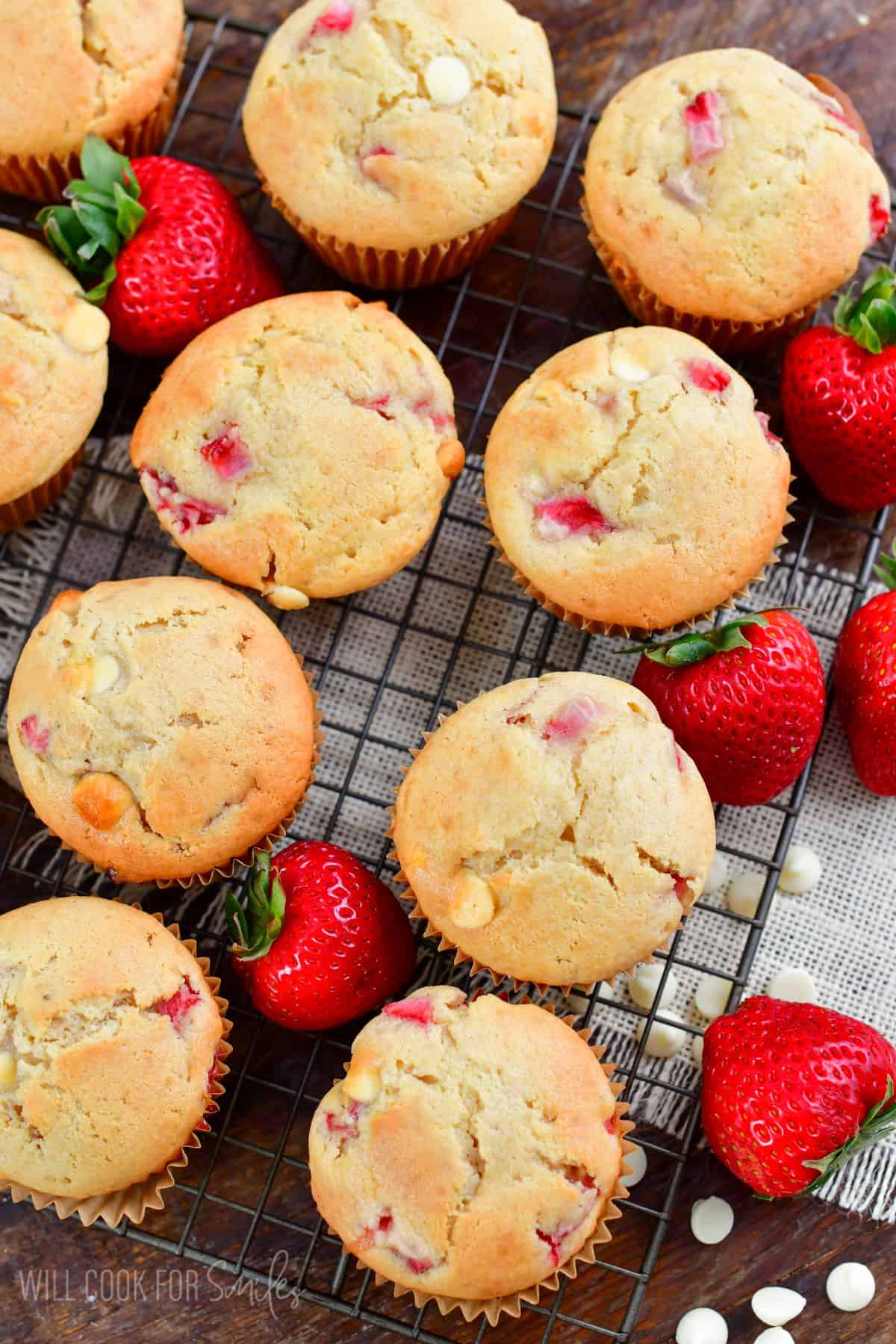 A bunch of white chocolate chip strawberry muffins on a cooking rack with strawberries around them.