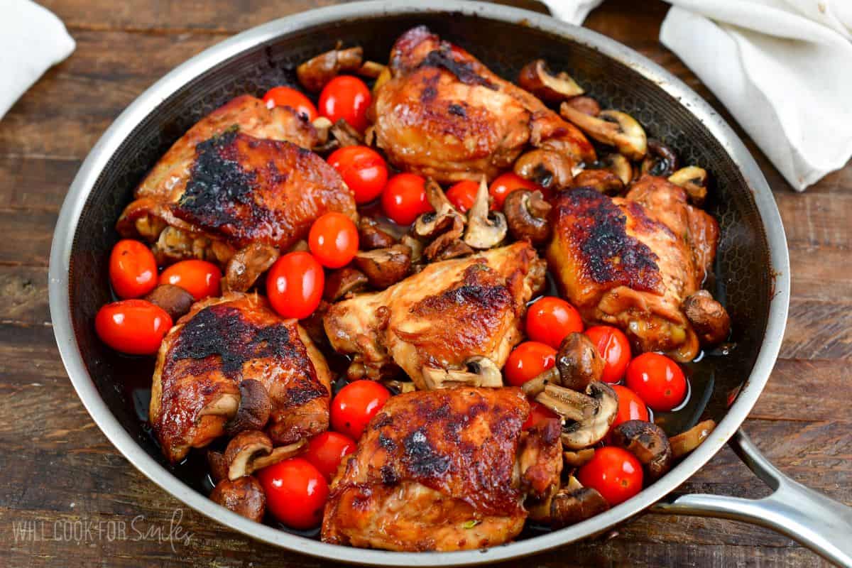 Balsamic baked chicken thighs in a pan with mushrooms and tomatoes in pan on a wood surface.