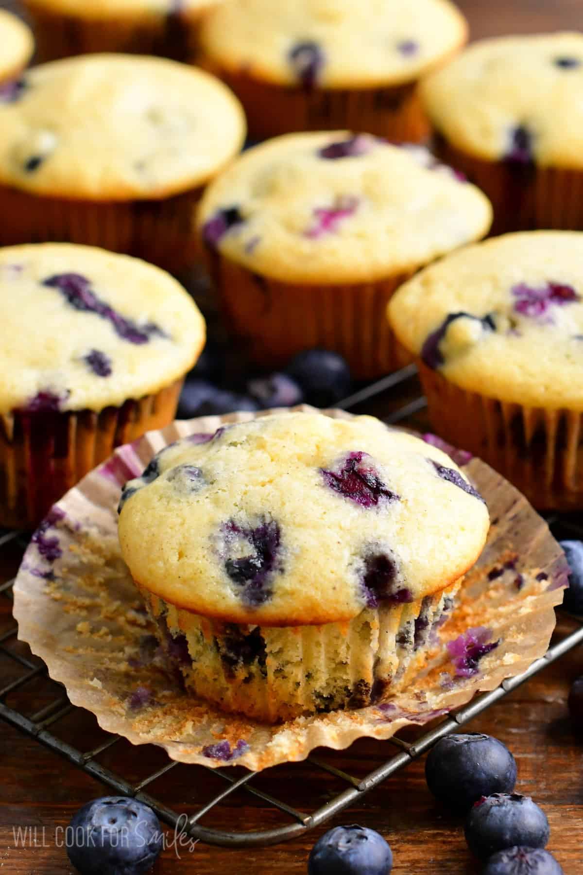 Blueberry muffins with the wrapper taken off on a wire rack with fresh blueberries around it.