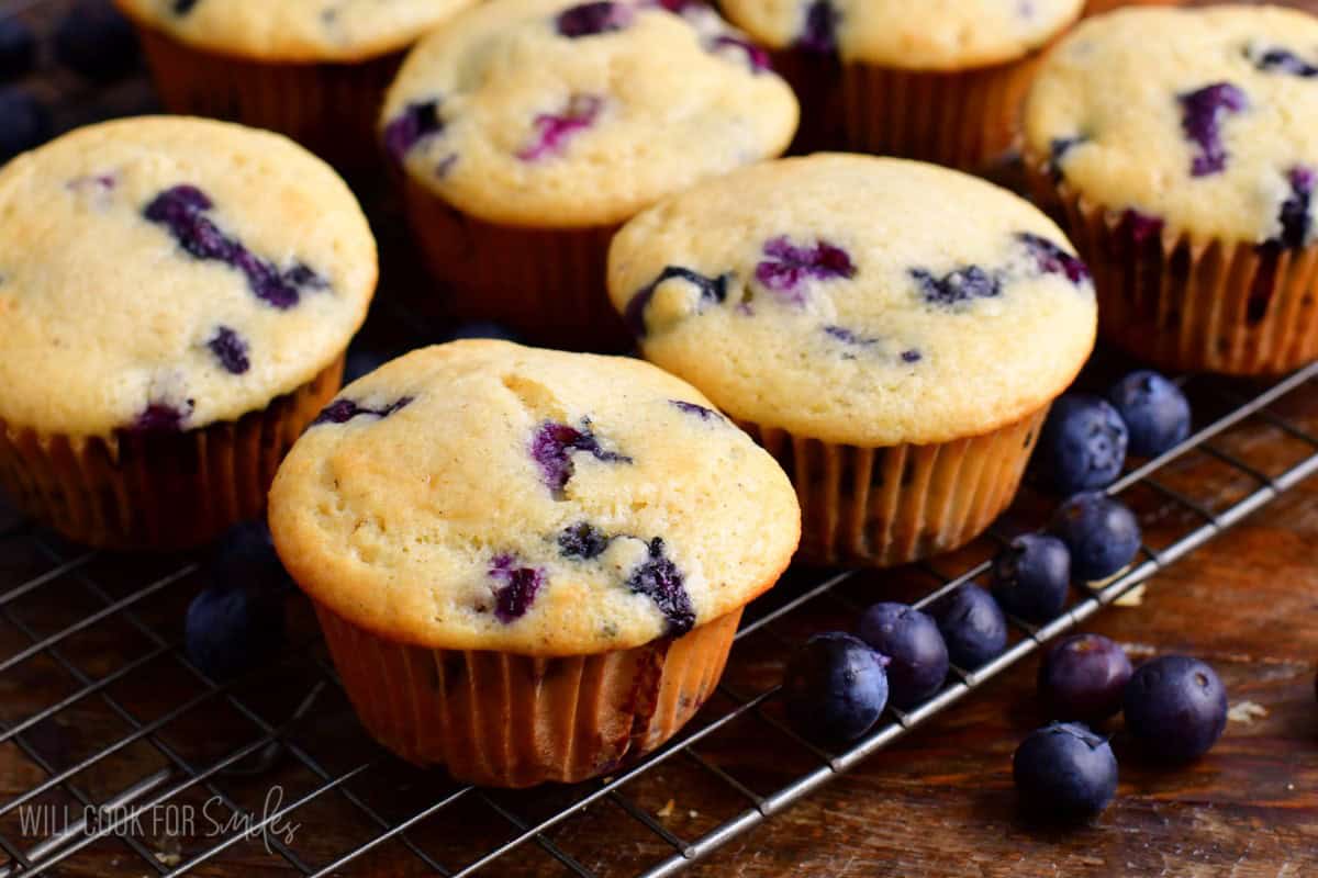 Several blueberry muffins on a wire cooling rack with blueberries around it.